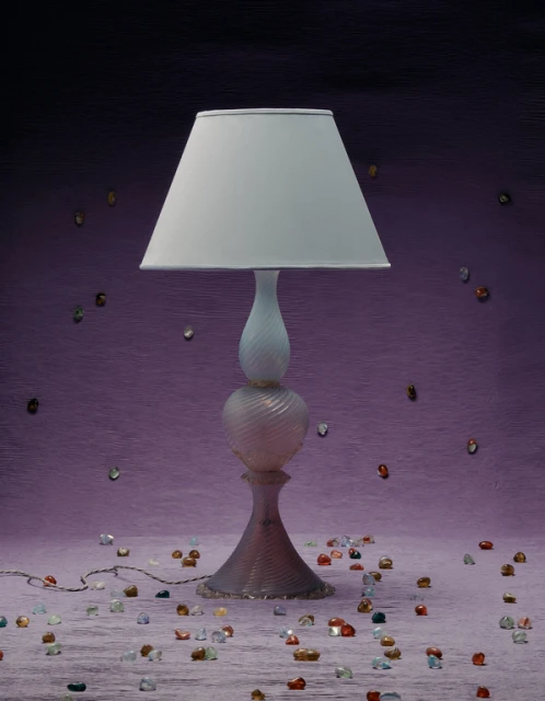 Illuminated glass table lamp with a white lampshade on a mauve textured background, surrounded by colorful decorative gems.