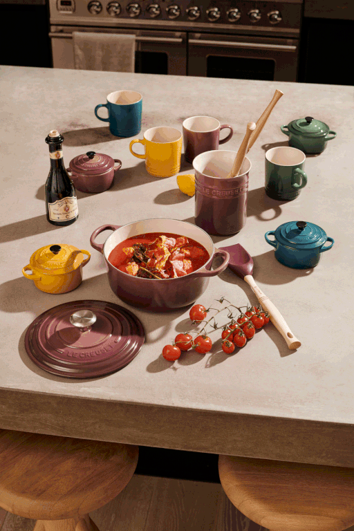 Overhead shot of a counter with colorful enameled cast iron cookware, mugs, serving utensils, tomatoes, and a small bottle of olive oil.