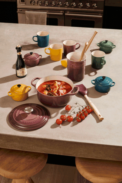 Overhead shot of a counter with colorful enameled cast iron cookware, mugs, serving utensils, tomatoes, and a small bottle of olive oil.
