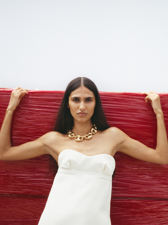 A woman poses in a white dress with a gold necklace, leaning against a bright red background, creating a striking fashion editorial image.
