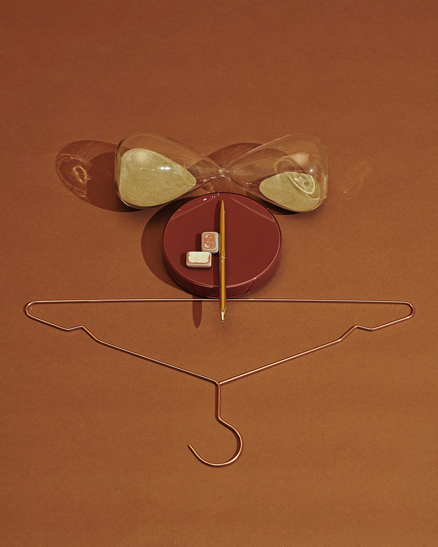 Still life photo with an hourglass, writing utensil, and hanger against a brown background. The composition is centrally framed.