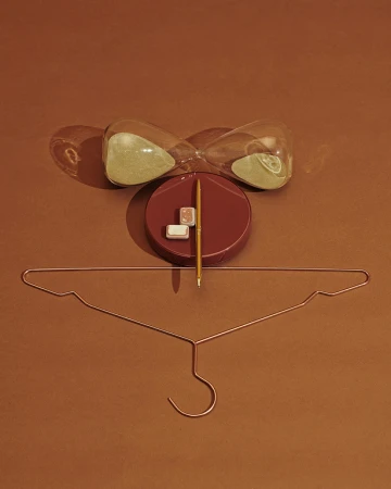 Still life photo with an hourglass, writing utensil, and hanger against a brown background. The composition is centrally framed.