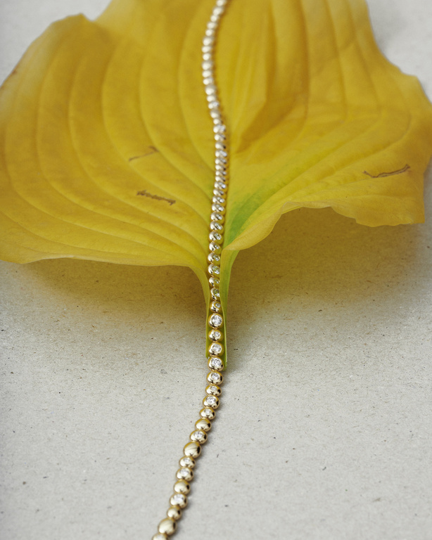 Close-up of a gold and diamond bracelet draped on a bright yellow leaf against a neutral grey surface, studio lighting.