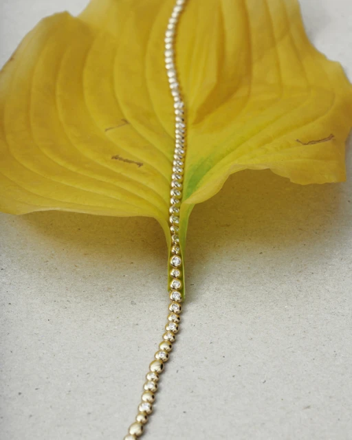 Close-up of a gold and diamond bracelet draped on a bright yellow leaf against a neutral grey surface, studio lighting.