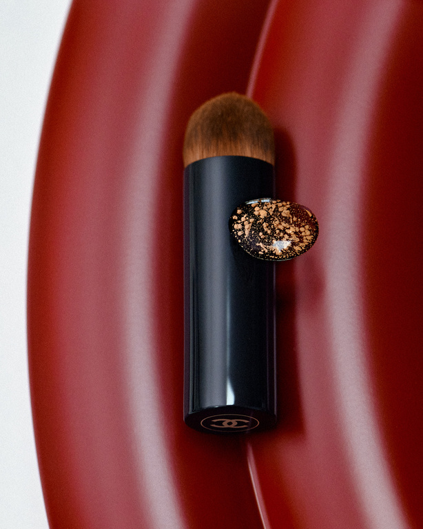 Chanel makeup brush with black handle and brown bristles, displayed against a rich, textured red surface. Beauty and cosmetic elegance.
