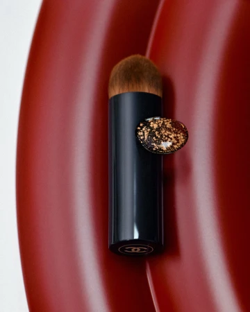 Chanel makeup brush with black handle and brown bristles, displayed against a rich, textured red surface. Beauty and cosmetic elegance.