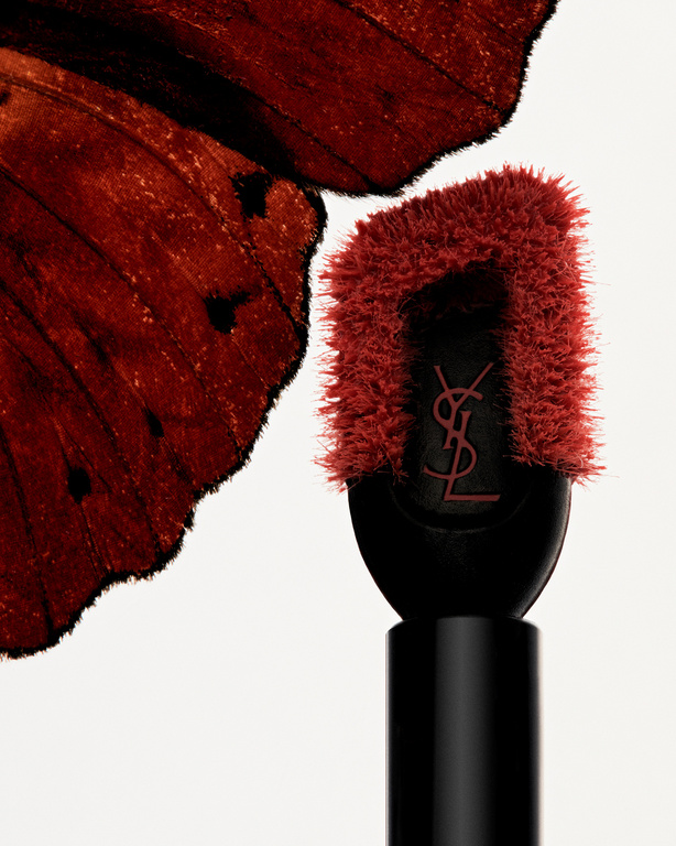 Close-up studio shot of YSL lipstick applicator and red butterfly wing against white. Beauty product and nature-inspired textures.