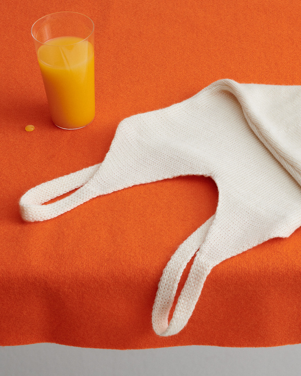 Still life shot of a glass of orange juice and a cream-colored knitted tote bag on an orange background, creating a vibrant and simple composition.