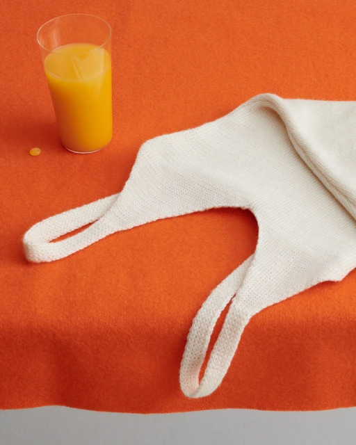 Still life shot of a glass of orange juice and a cream-colored knitted tote bag on an orange background, creating a vibrant and simple composition.