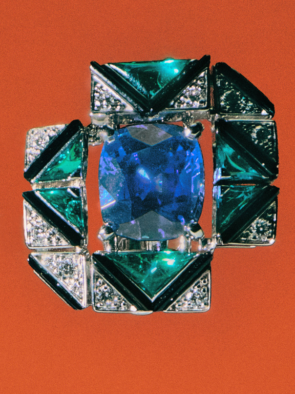 Close-up shot of a silver brooch featuring a large, cushion-cut blue gemstone surrounded by green and black enamel with diamond accents.