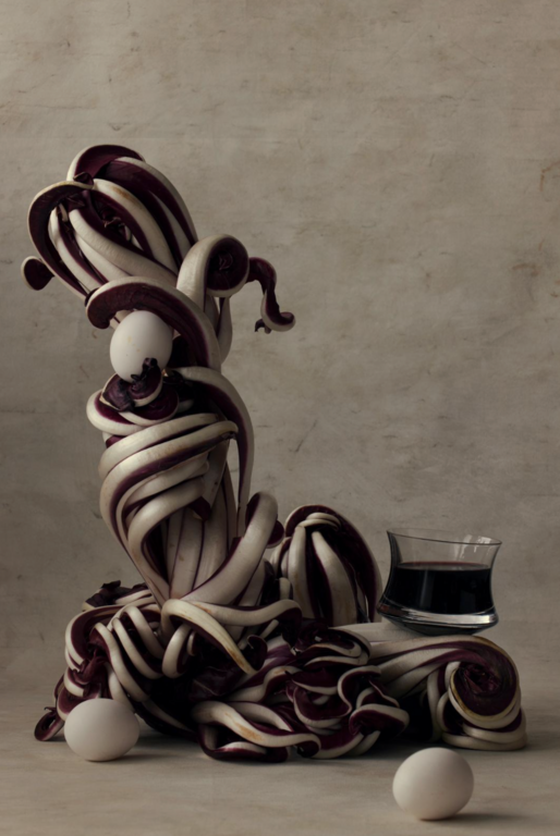 Still life photograph of radicchio arranged vertically with white eggs and a glass of dark red liquid against a textured backdrop.