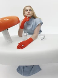 Woman in blue dress wearing bright orange gloves and rings, seated at a white table with an orange lamp