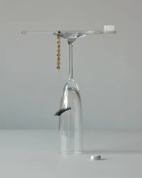 An inverted wine glass balanced with a toothbrush, gold chain, false eyelash, and tablet on a light grey seamless backdrop.