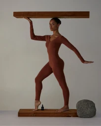 Woman in terracotta bodysuit balancing a wooden beam with a small bottle and stone on a grey seamless backdrop