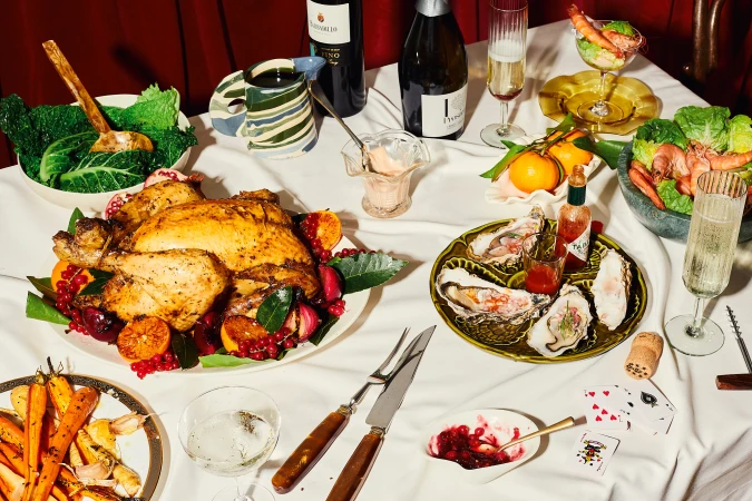 Overhead shot of a festive dining table adorned with a roasted chicken, oysters, wine, champagne, and various appetizers, creating a rich holiday feast.