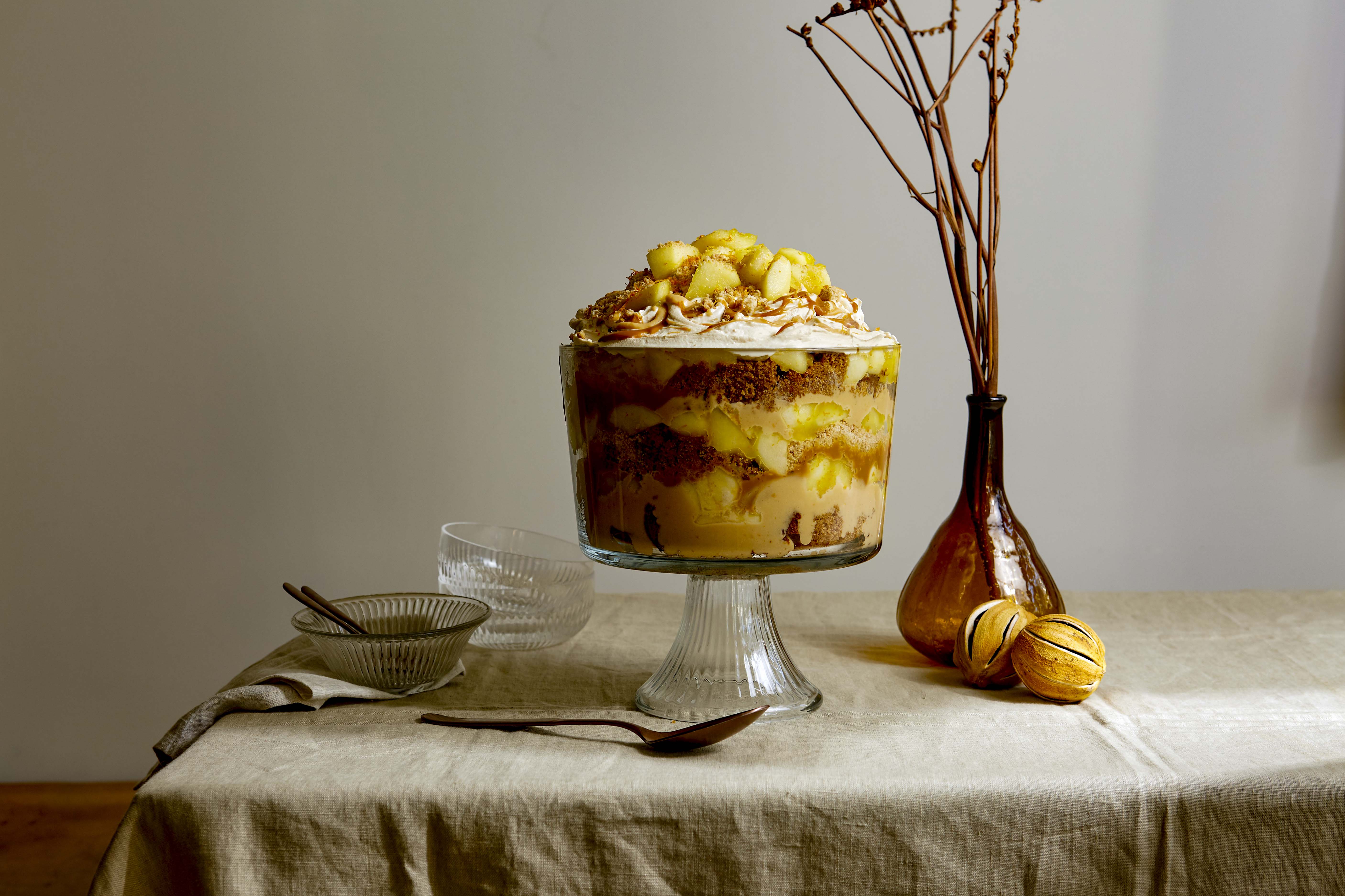A trifle dessert sits in a glass stand on a linen-covered table, with small bowls and dried oranges, in an indoor setting with soft lighting.
