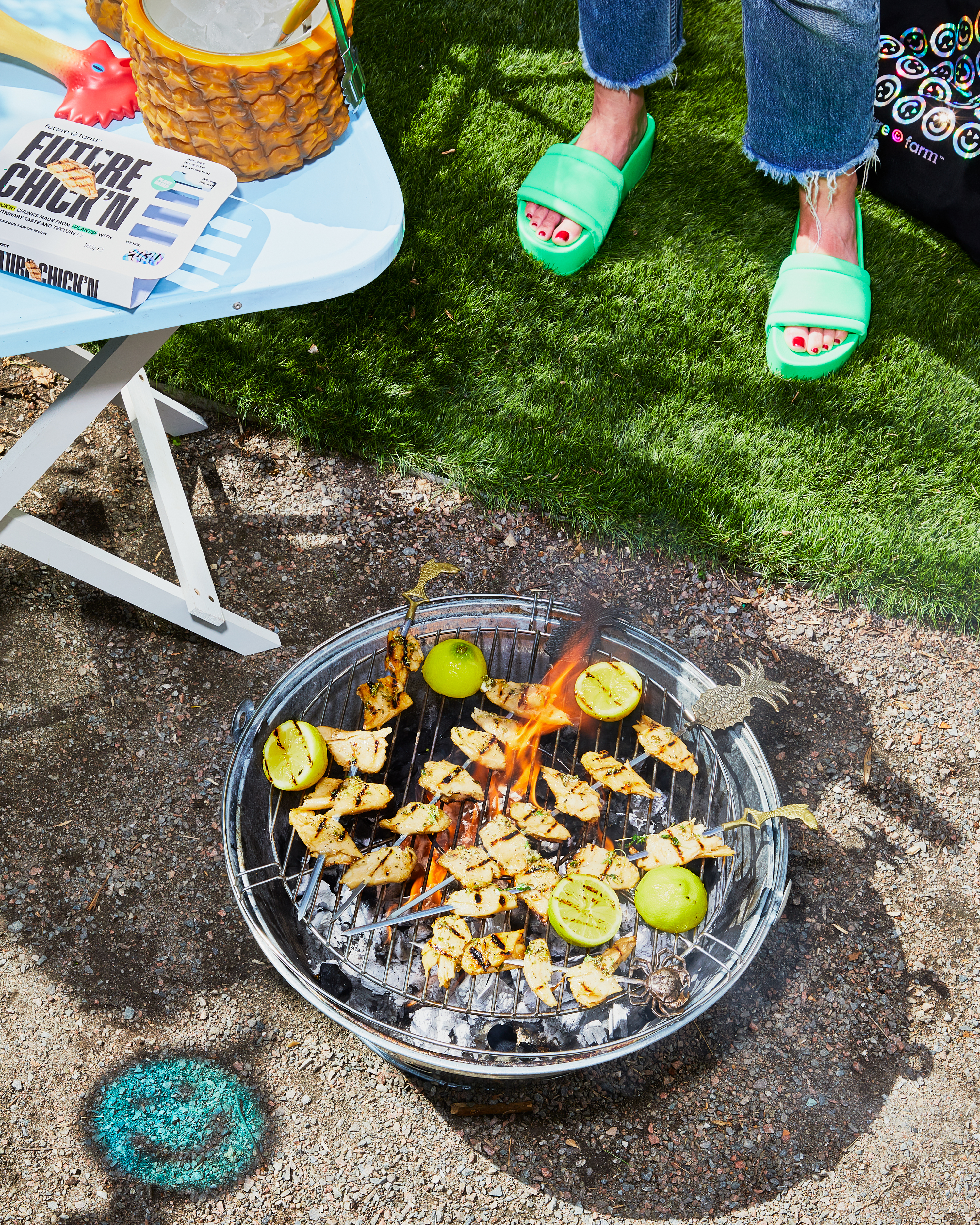 A metal grill with chicken and limes being cooked on a portable charcoal grill, with a table and a woman's legs and shoes in the shot.