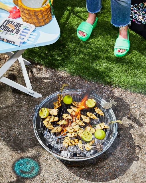 A metal grill with chicken and limes being cooked on a portable charcoal grill, with a table and a woman's legs and shoes in the shot.