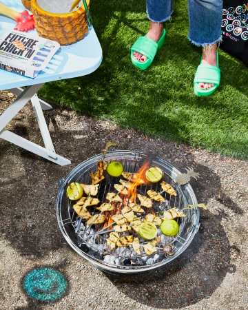A metal grill with chicken and limes being cooked on a portable charcoal grill, with a table and a woman's legs and shoes in the shot.