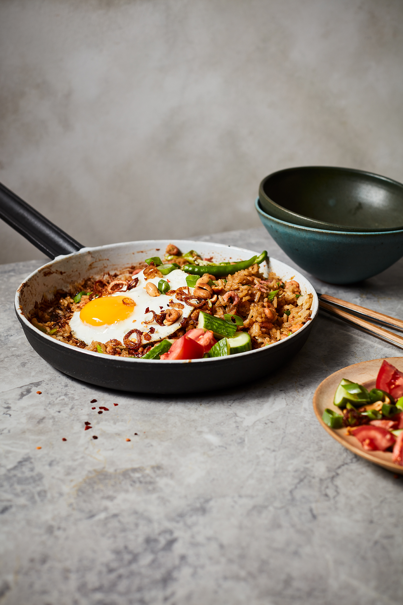 Overhead shot of fried rice with a fried egg in a skillet, accompanied by bowls, wooden chopsticks, and salad plates on a gray surface.
