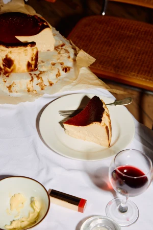 Slice of Basque burnt cheesecake on a plate with a fork, glass of red wine, lipstick, and a lit candle on a white tablecloth.