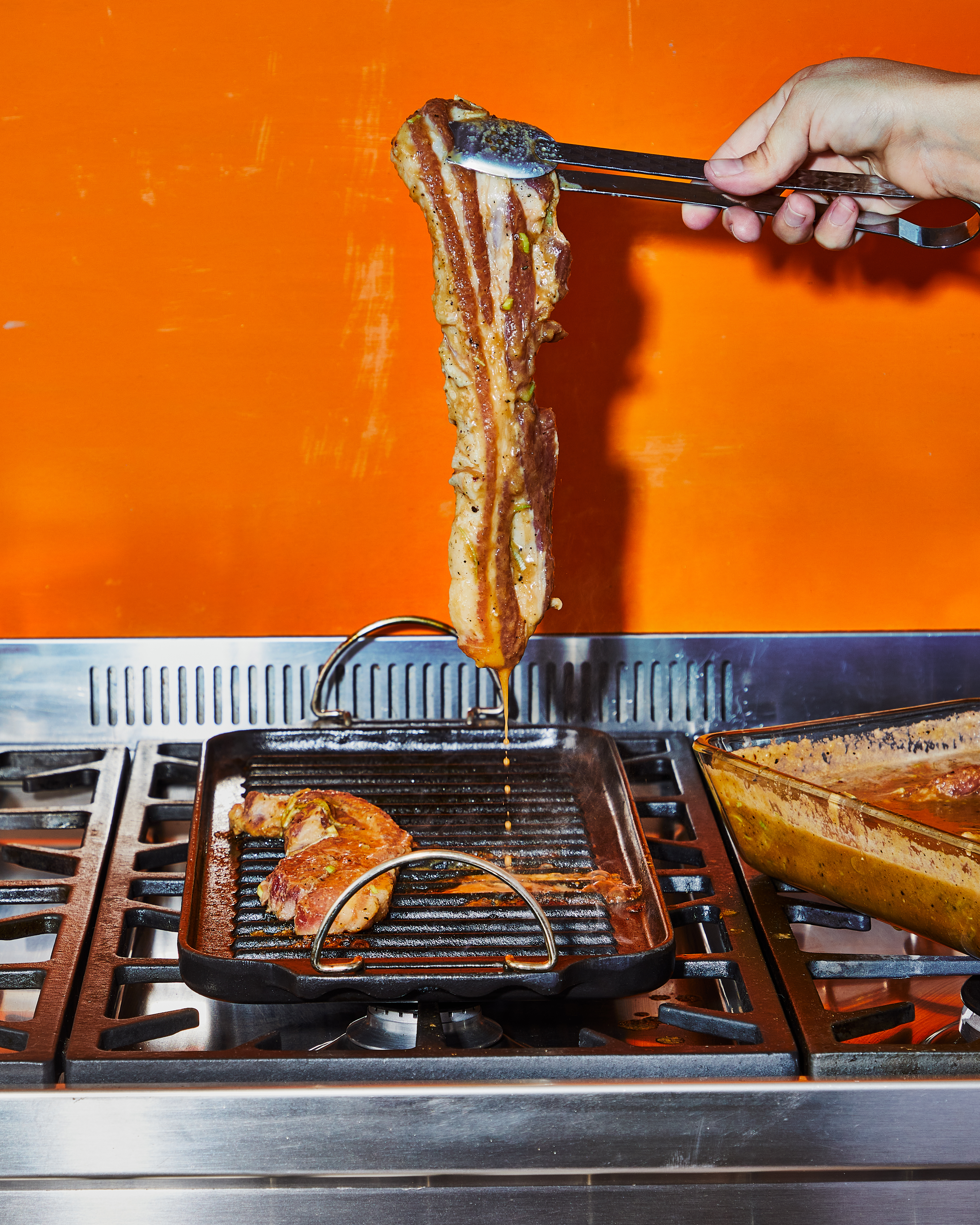 Pork belly being cooked on a stovetop grill, held up with metal tongs. Orange wall in the background. Food preparation in progress.