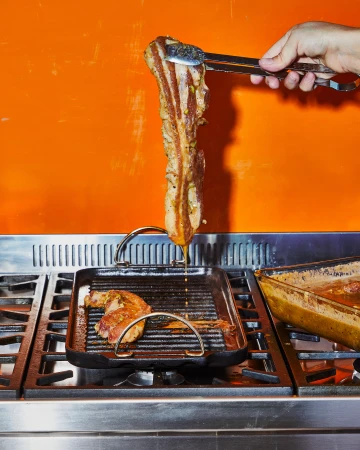 Pork belly being cooked on a stovetop grill, held up with metal tongs. Orange wall in the background. Food preparation in progress.