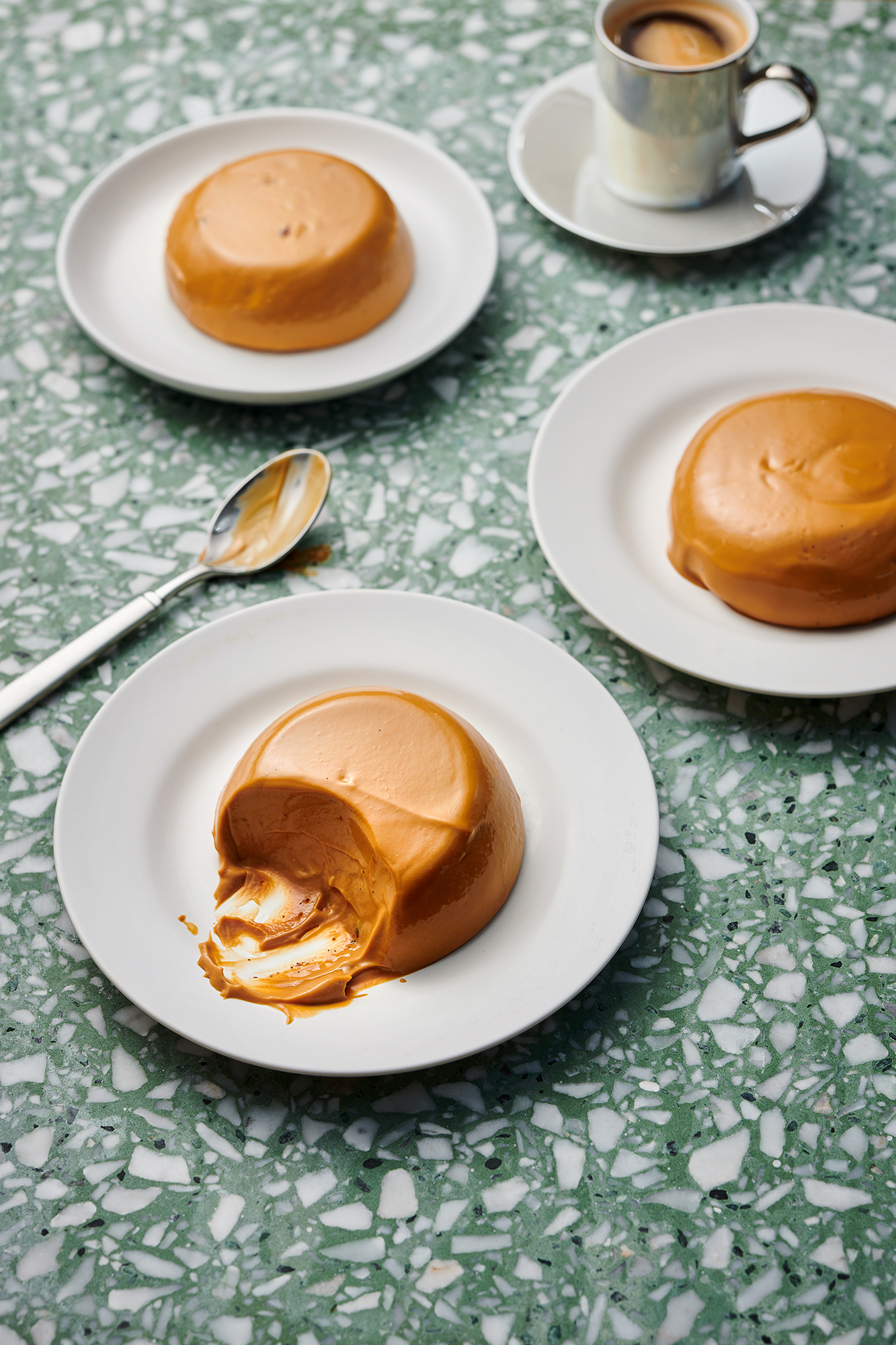 Overhead shot of three caramel desserts on white plates, coffee cup, silver spoon on green terrazzo surface; food photography.