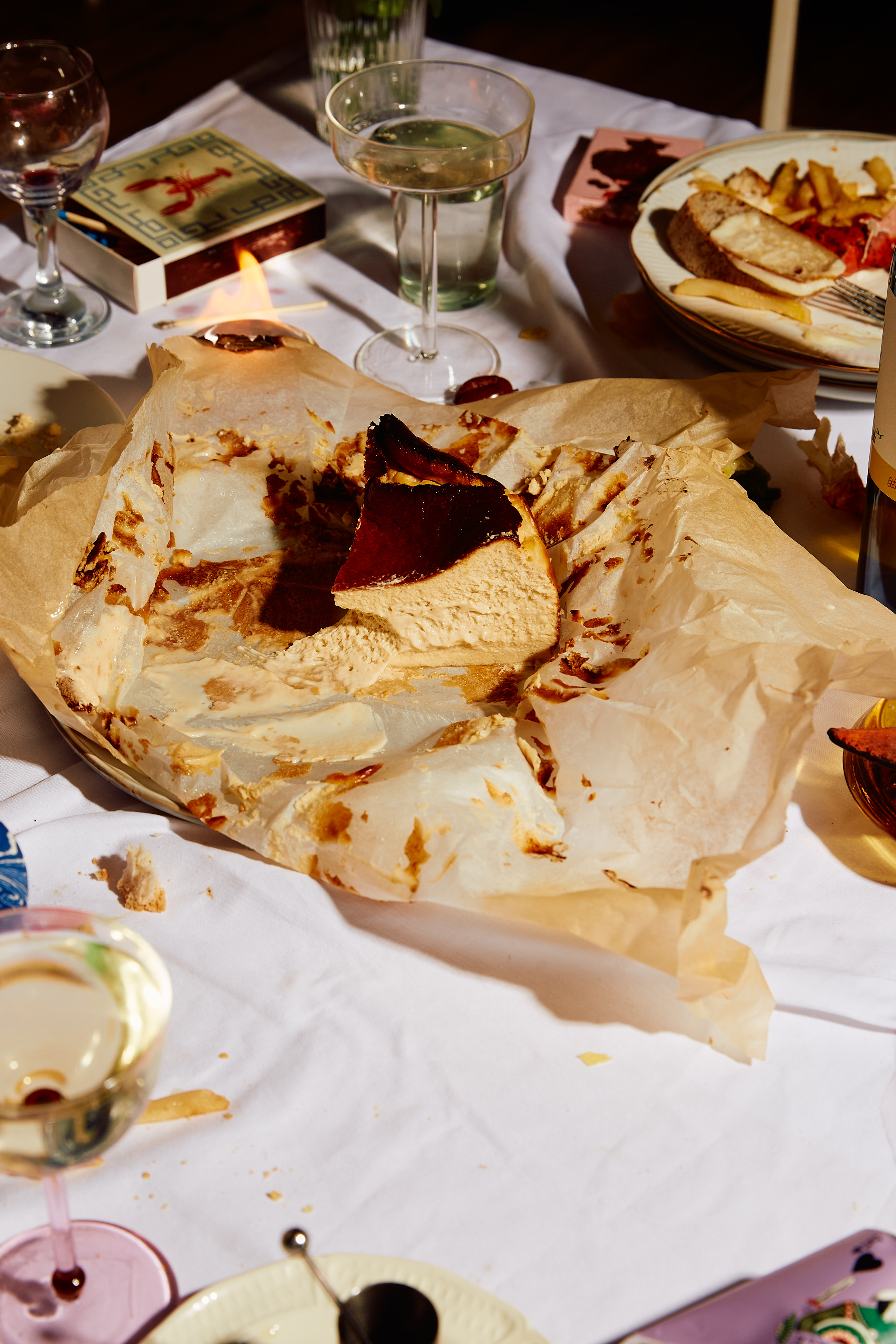 Slice of Basque burnt cheesecake on parchment paper on a white tablecloth, with wine glasses, plates, matches, and candle.