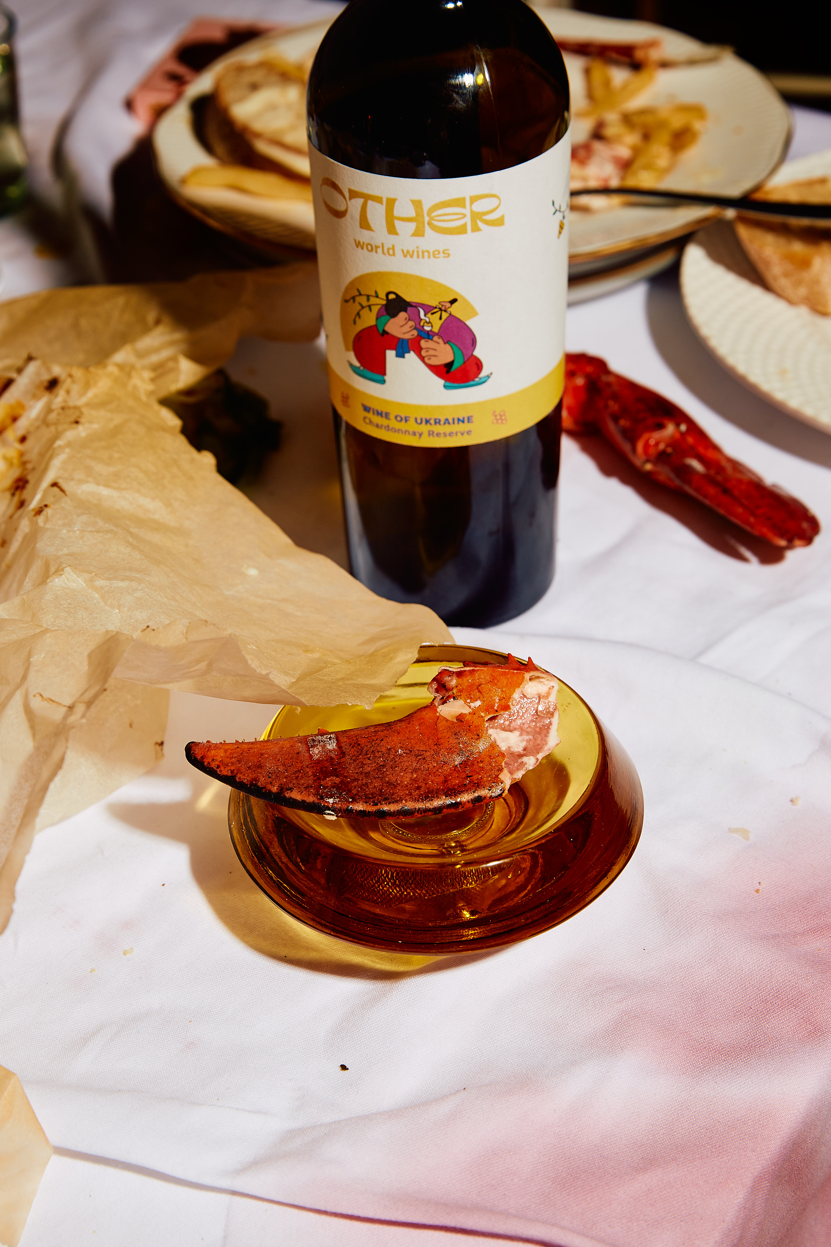 Wine bottle with an 'OTHER world wines' label, paired with a glass featuring a lobster claw, on a white tablecloth with food remnants.