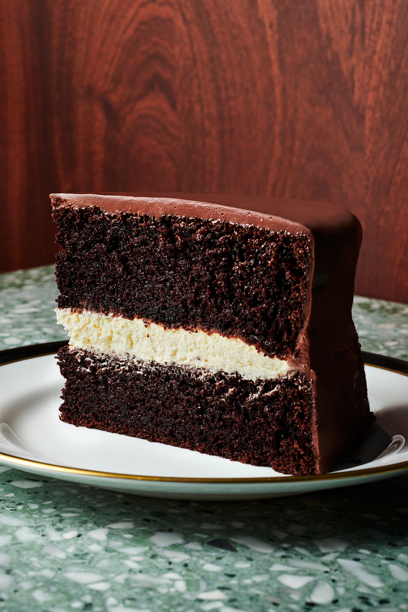 Close-up of a slice of chocolate cake with cream filling on a white plate with gold trim, set against a wooden background.