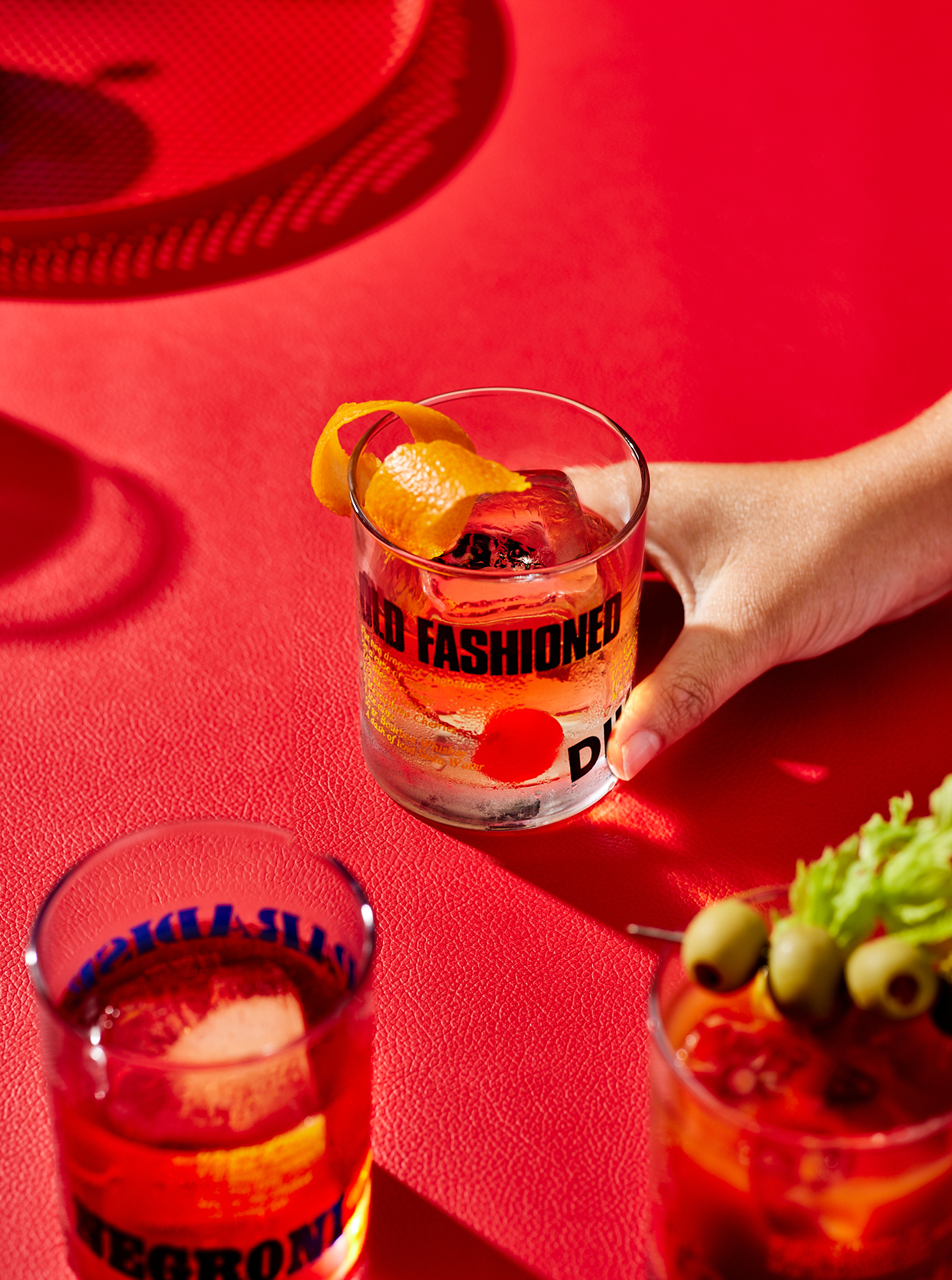 Three cocktails are arranged on a red surface: an Old Fashioned, a Negroni, and a Bloody Mary, with a hand holding the Old Fashioned.