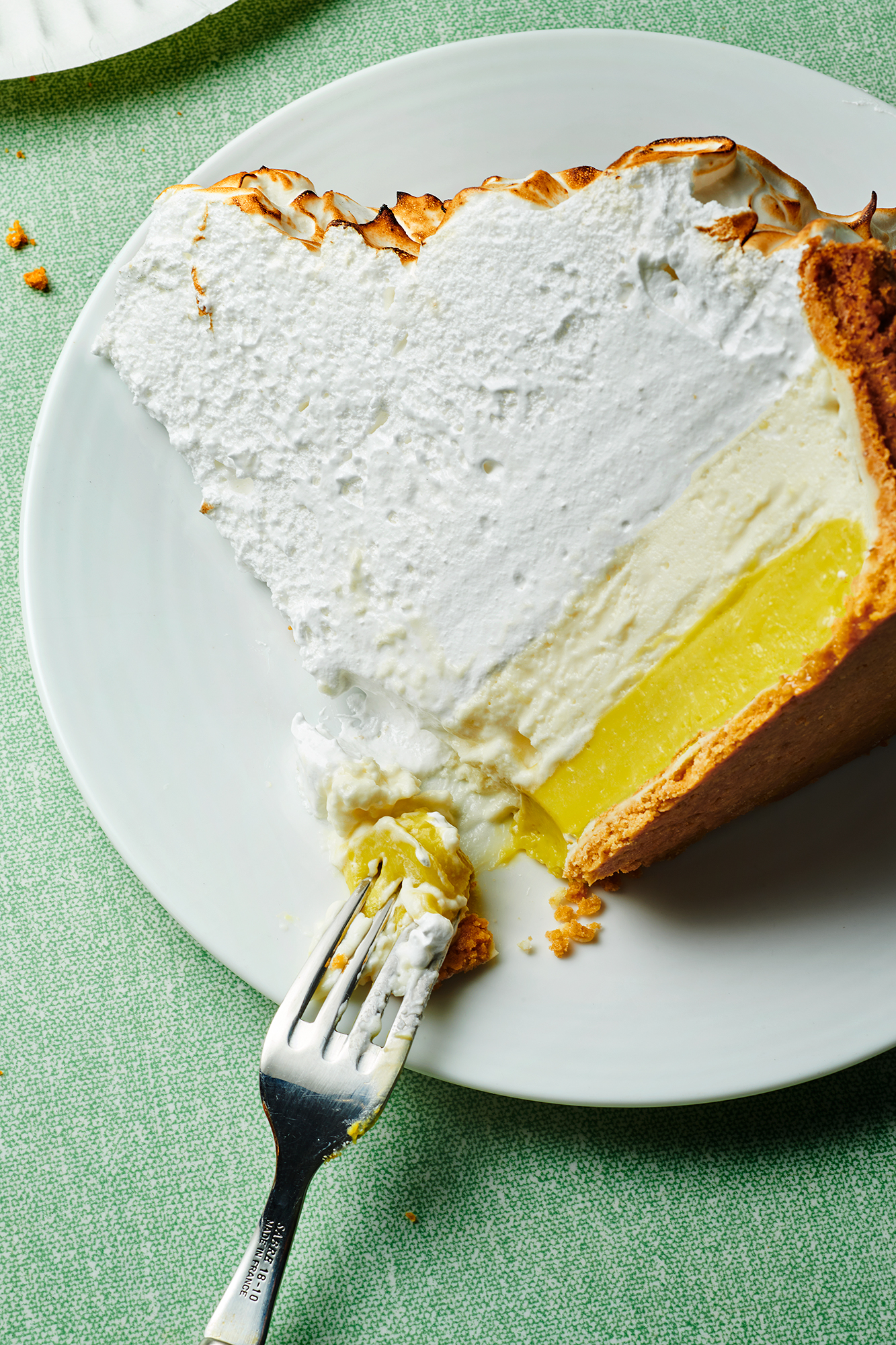 Overhead shot of a slice of lemon meringue pie on a white plate, served with a fork on a textured green tablecloth.