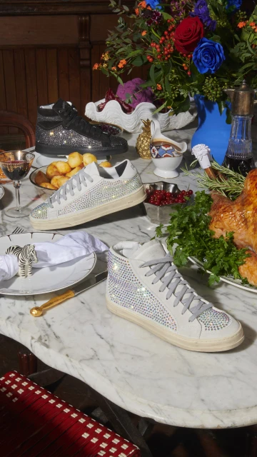 Elevated product shot showcasing black and white embellished sneakers arranged on a white marble table with dining and floral decor.