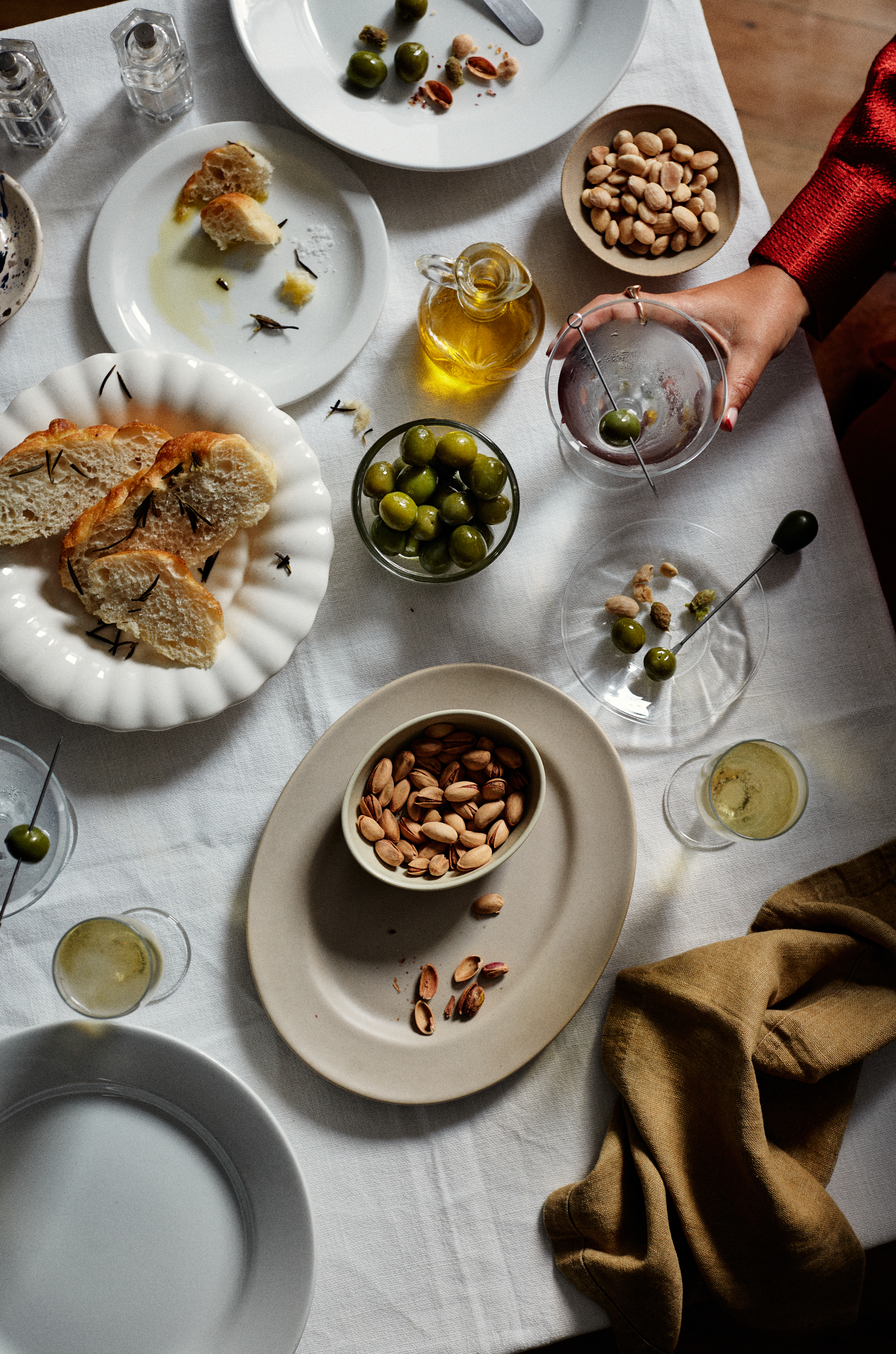 Overhead shot of an off-white linen tablecloth laden with plates of bread, olives, almonds, and drinks, creating an inviting food still life.