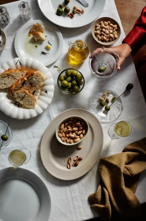 Overhead shot of an off-white linen tablecloth laden with plates of bread, olives, almonds, and drinks, creating an inviting food still life.