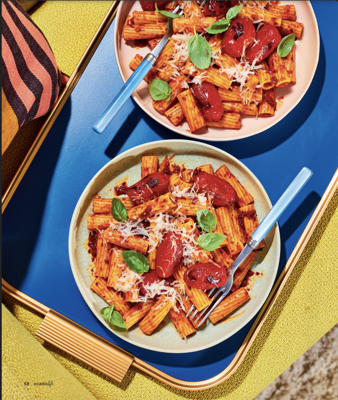 Overhead shot of two plates of rigatoni pasta with tomato sauce, roasted tomatoes, basil, and grated cheese on a colorful table.