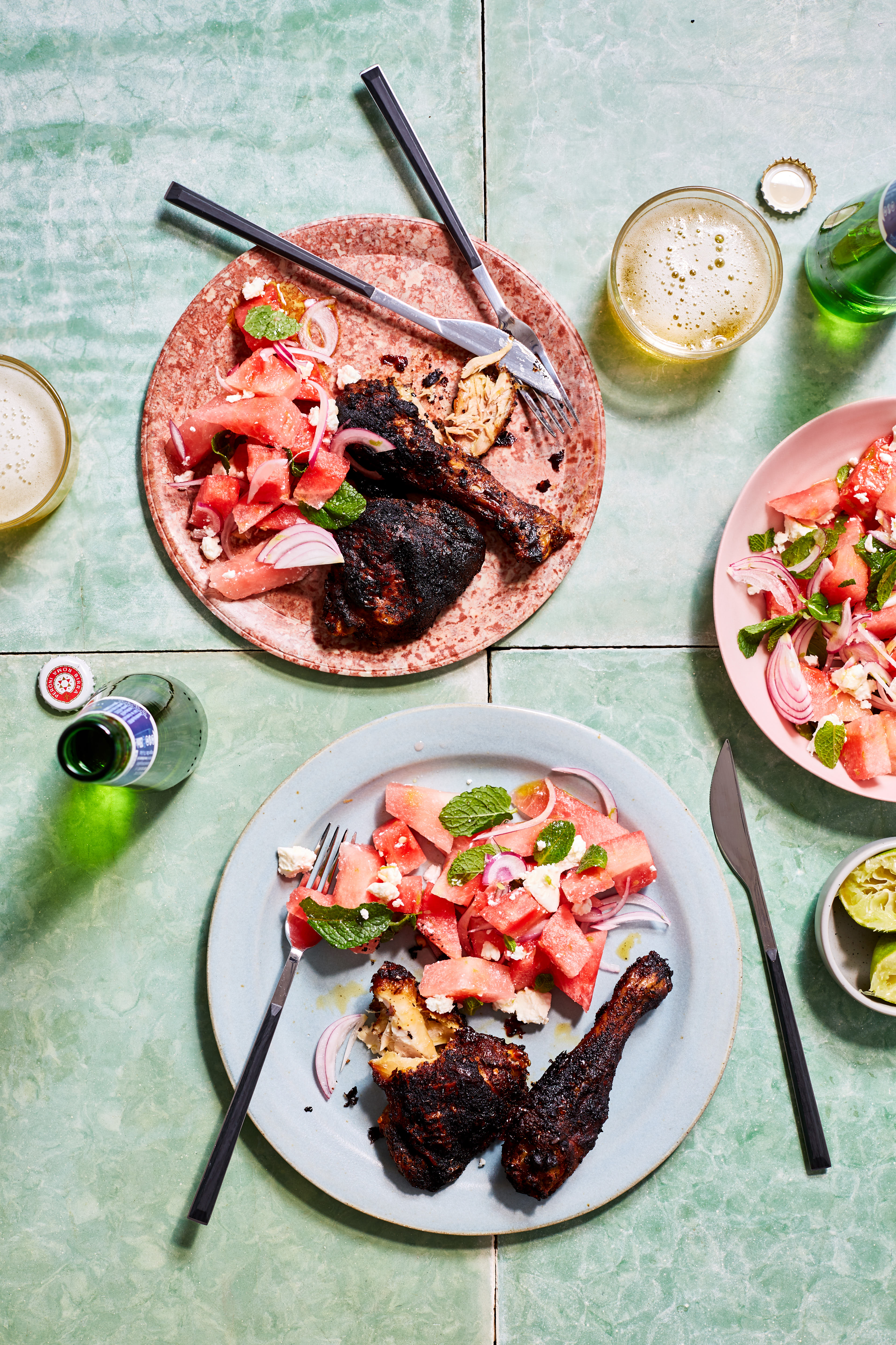 Overhead shot of two plates of grilled chicken, watermelon salad, beer, and limes arranged on a green tiled surface.