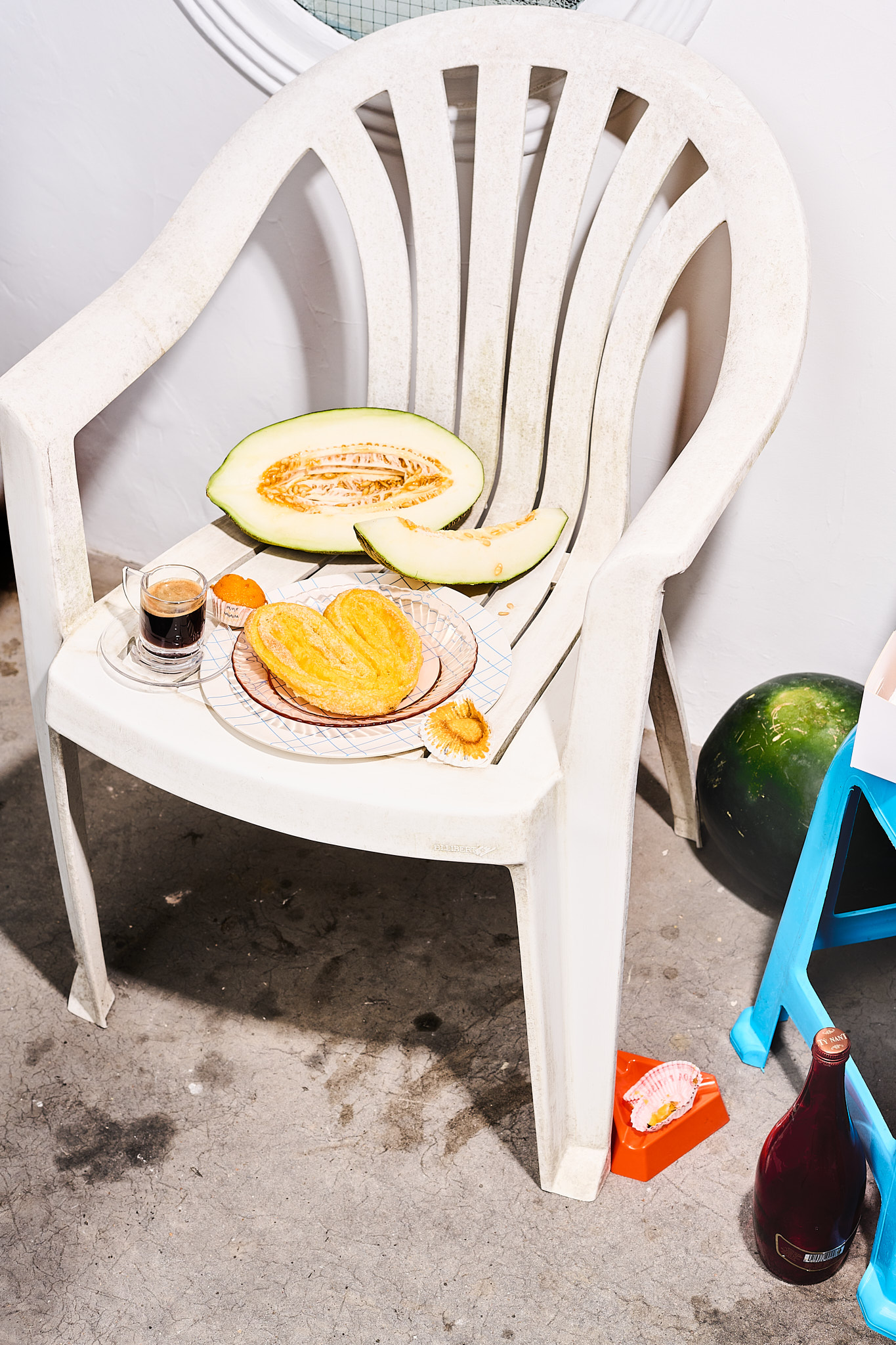 Still life of cantaloupe melon, pastry, and espresso on a white plastic chair, outdoors with a cement floor and white wall backdrop.