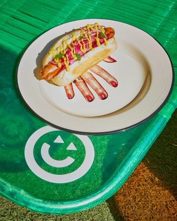Overhead shot of a hot dog with mustard and toppings on a plate, resting on a green tray with a smiley face graphic.