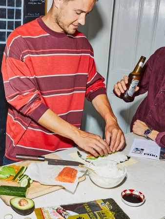 Two people are making sushi at a table with ingredients like seaweed, rice, avocado, and salmon; one person is holding a beer bottle.