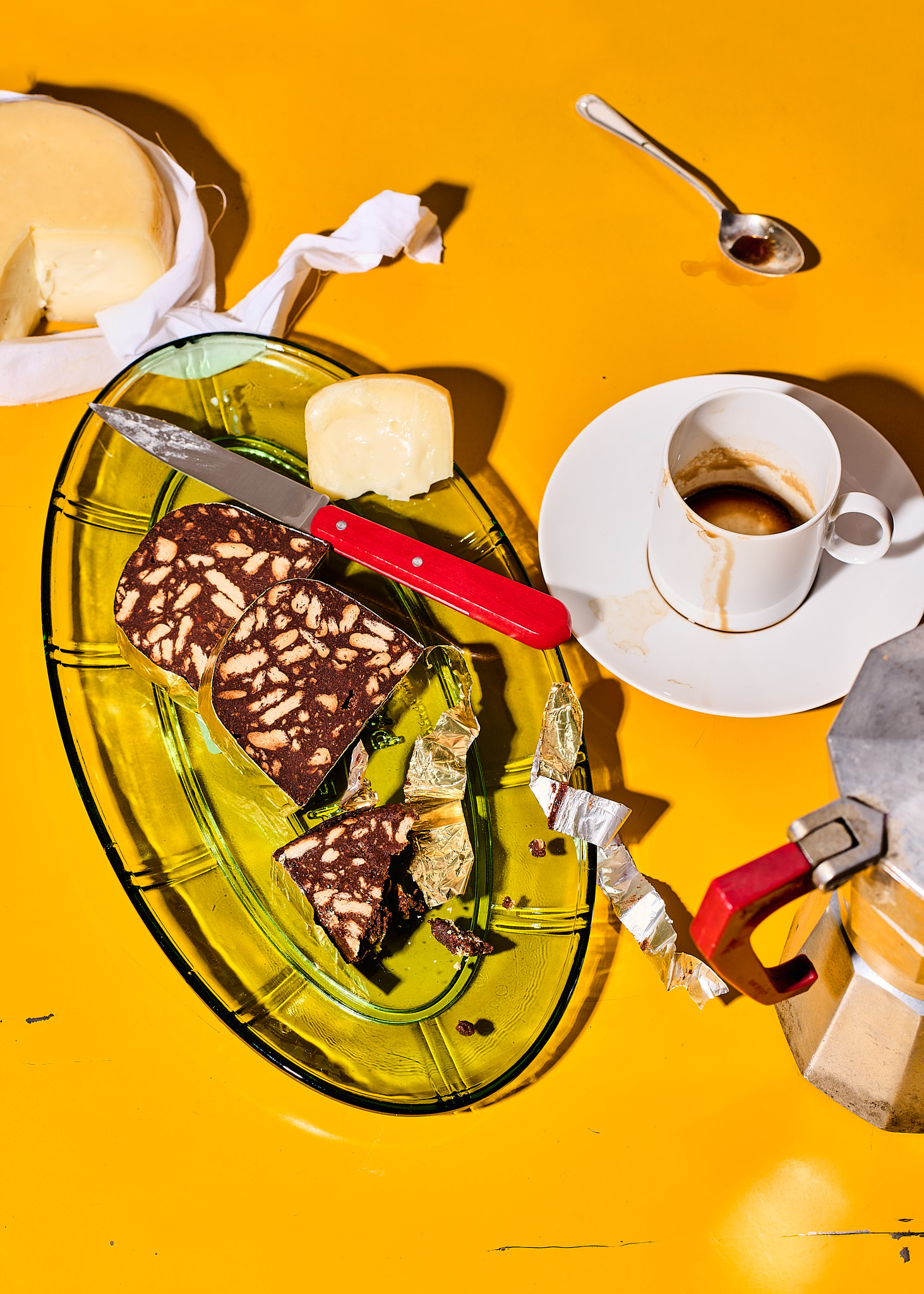 Overhead shot of a yellow table with cheese, chocolate salami, a vintage espresso maker, and a coffee cup, showcasing food photography.