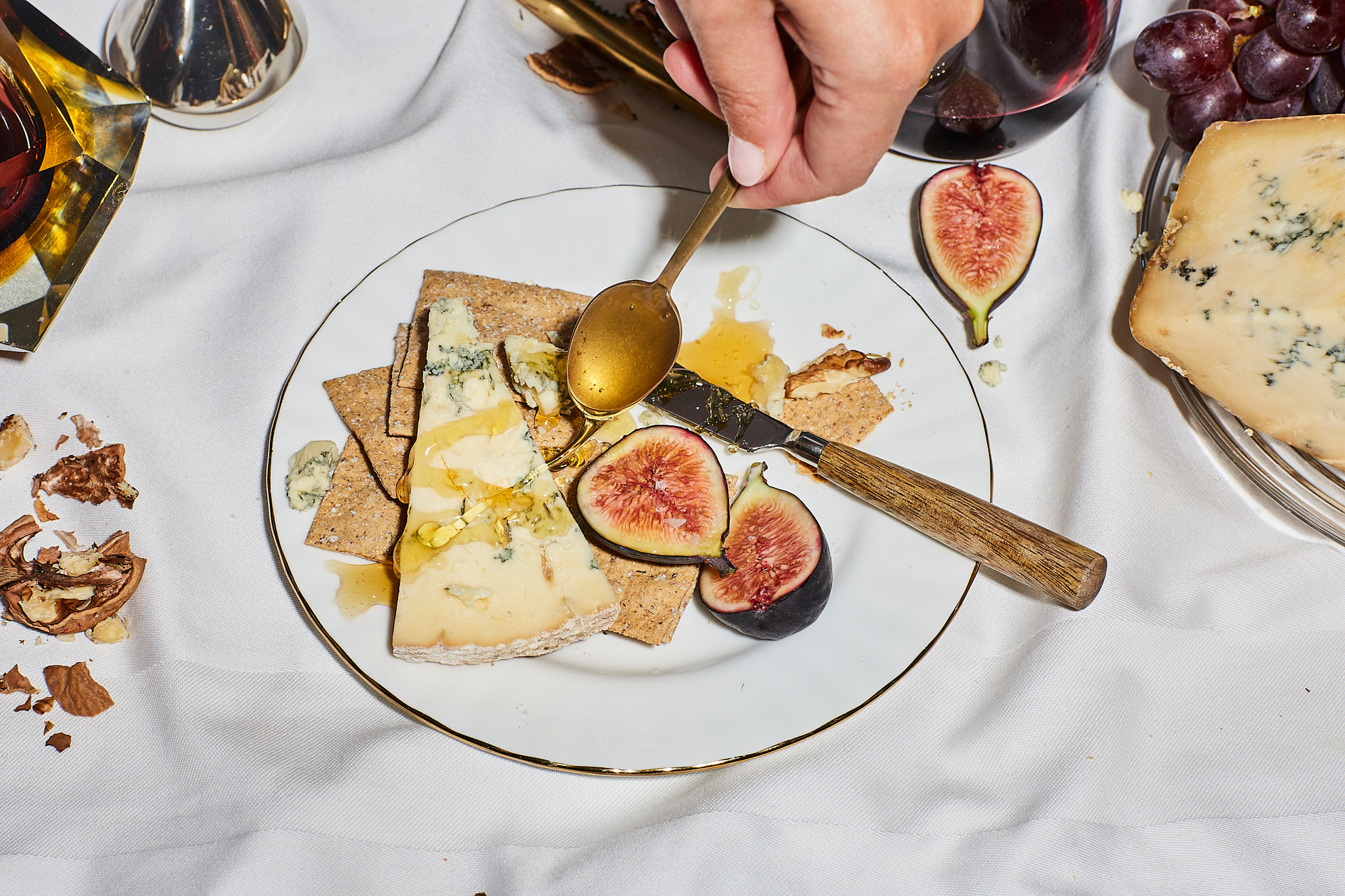 Overhead shot of a plate with blue cheese, crackers, figs, and honey being drizzled by a hand holding a golden spoon. A glass of wine is nearby.