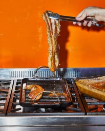 Hand holding metal tongs lifting a strip of cooked pork belly above a black ribbed grill pan in a kitchen