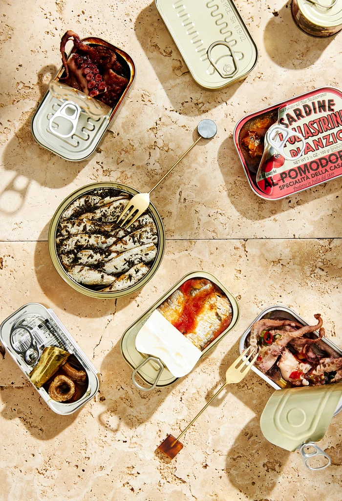 Top-down view of open canned seafood tins with sardines and octopus on a textured stone surface with small gold forks.