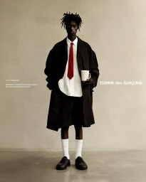 Black man wearing oversized black coat, white shirt, red tie, white socks, and holding a mug against a plain wall