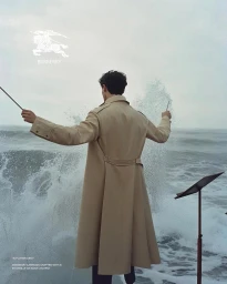 Man wearing a long tan trench coat conducting ocean waves with batons on a beach by Sybille de Saint Louvent