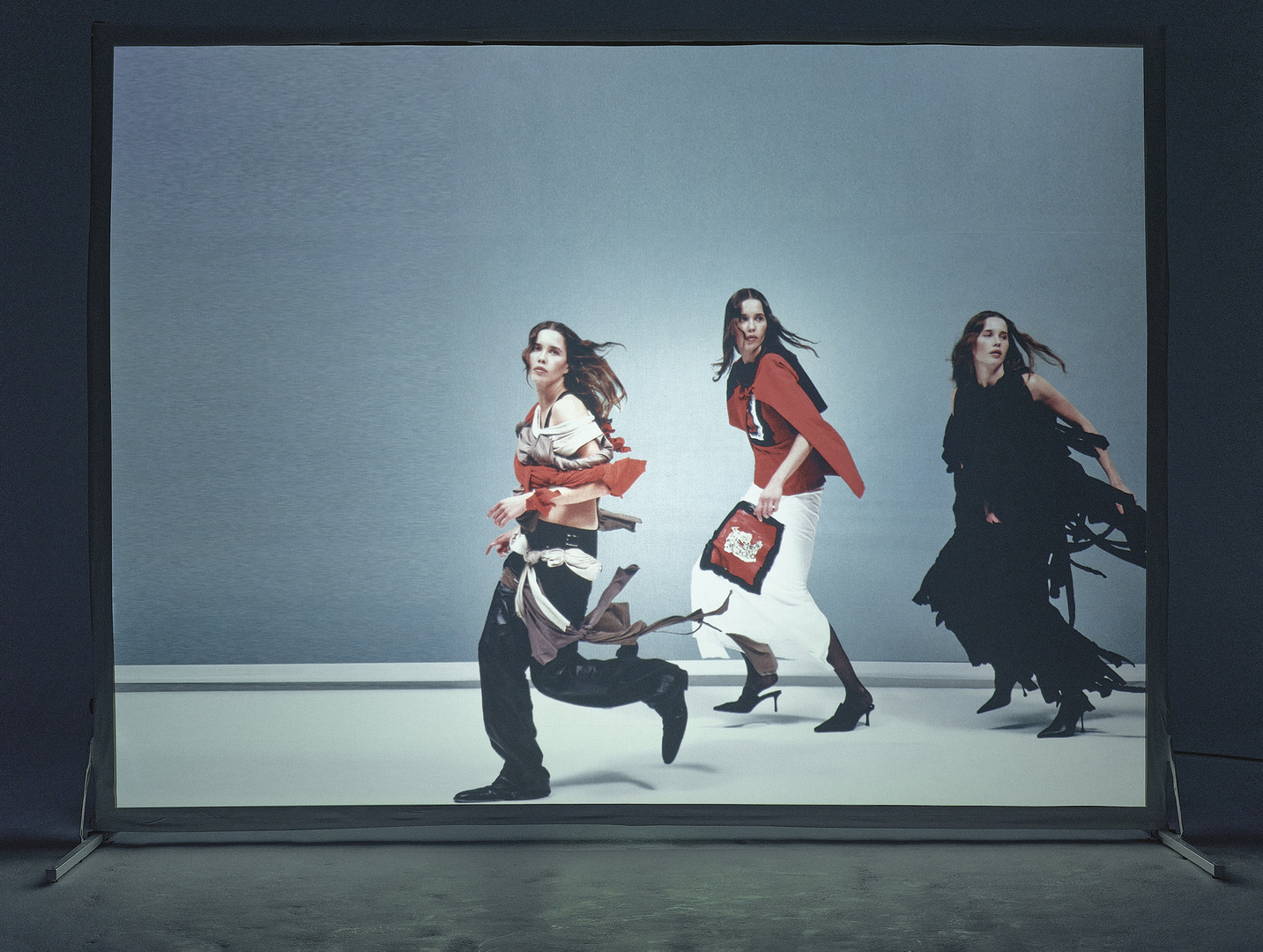 Three fashion models in motion wearing unique outfits in a studio. The image shows an action pose against a simple grey backdrop.