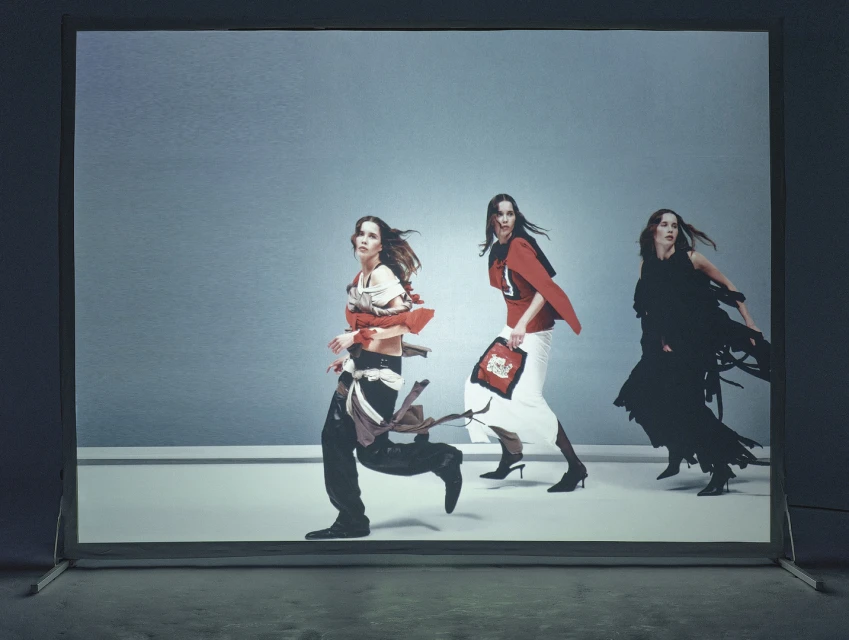Three fashion models in motion wearing unique outfits in a studio. The image shows an action pose against a simple grey backdrop.