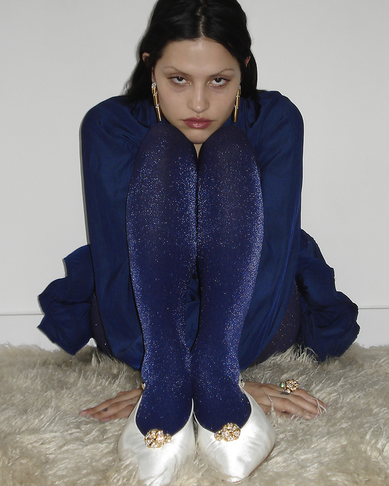 Portrait of a young woman with long dark hair, wearing a blue top, blue sparkly tights, jeweled ballet flats, and gold earrings.